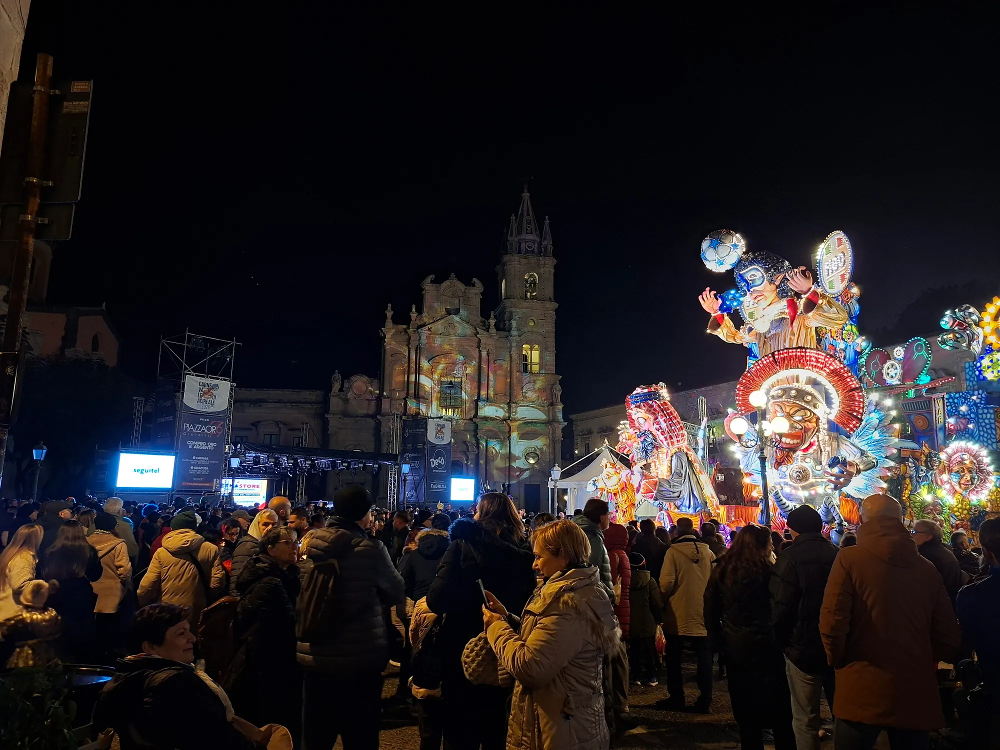 A town square at night. A tall baroque church front with a pierced bell tower is lit with coloured projections; a stage with screens and banners stands to the left, and a crowd in winter coats fills the foreground. At the right edge, an illuminated float carrying huge grotesque moulded faces rises above the heads.