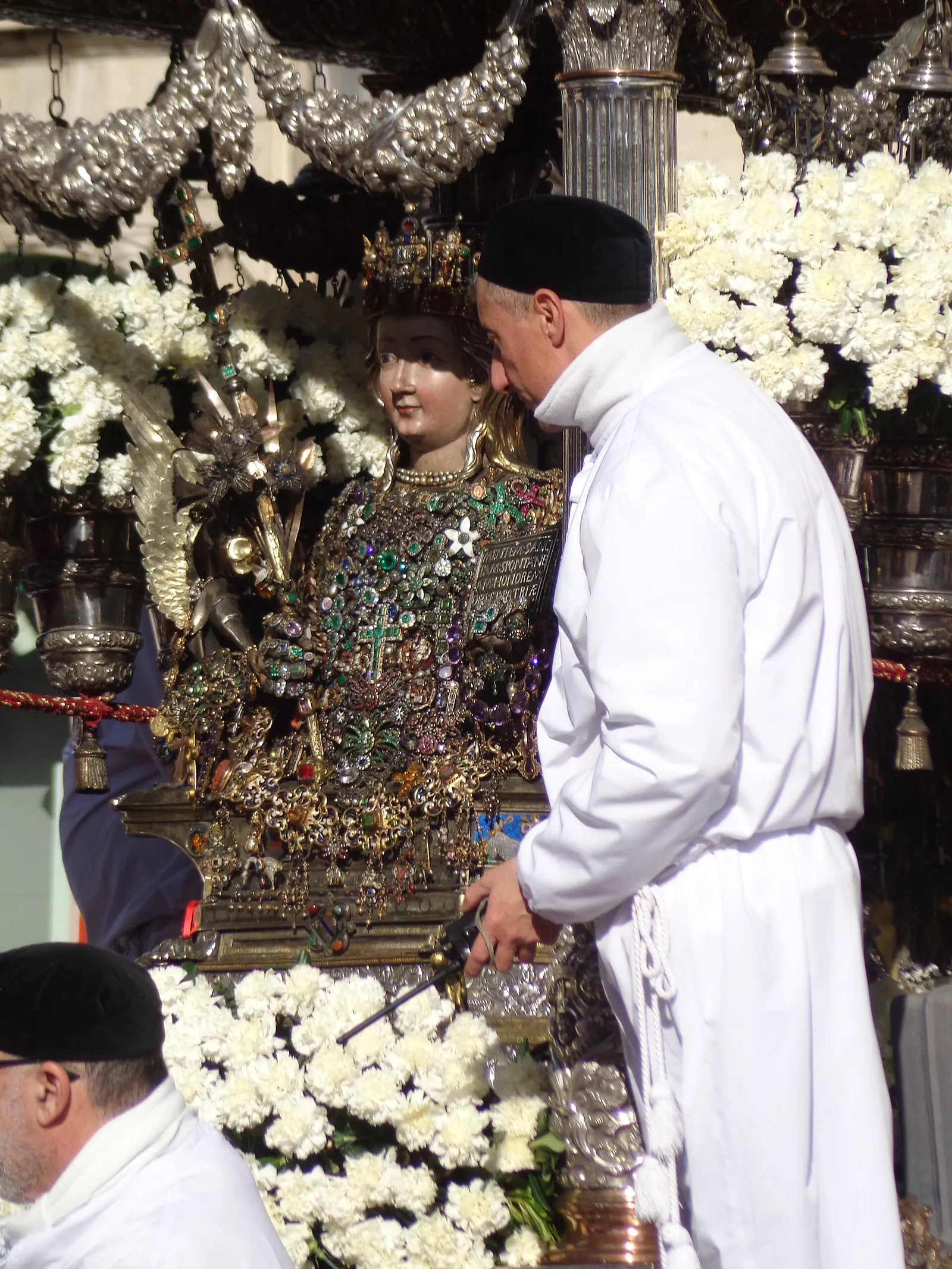 Close on a reliquary bust: a painted face under a tall jewelled crown, a pearl necklace, and a breastplate so densely covered in brooches, crosses, stars and coloured stones that little of the metal beneath is visible. A small inscribed plate sits on the shoulder. A man in a white smock and black skullcap leans in beside it, and white carnations bank up below.