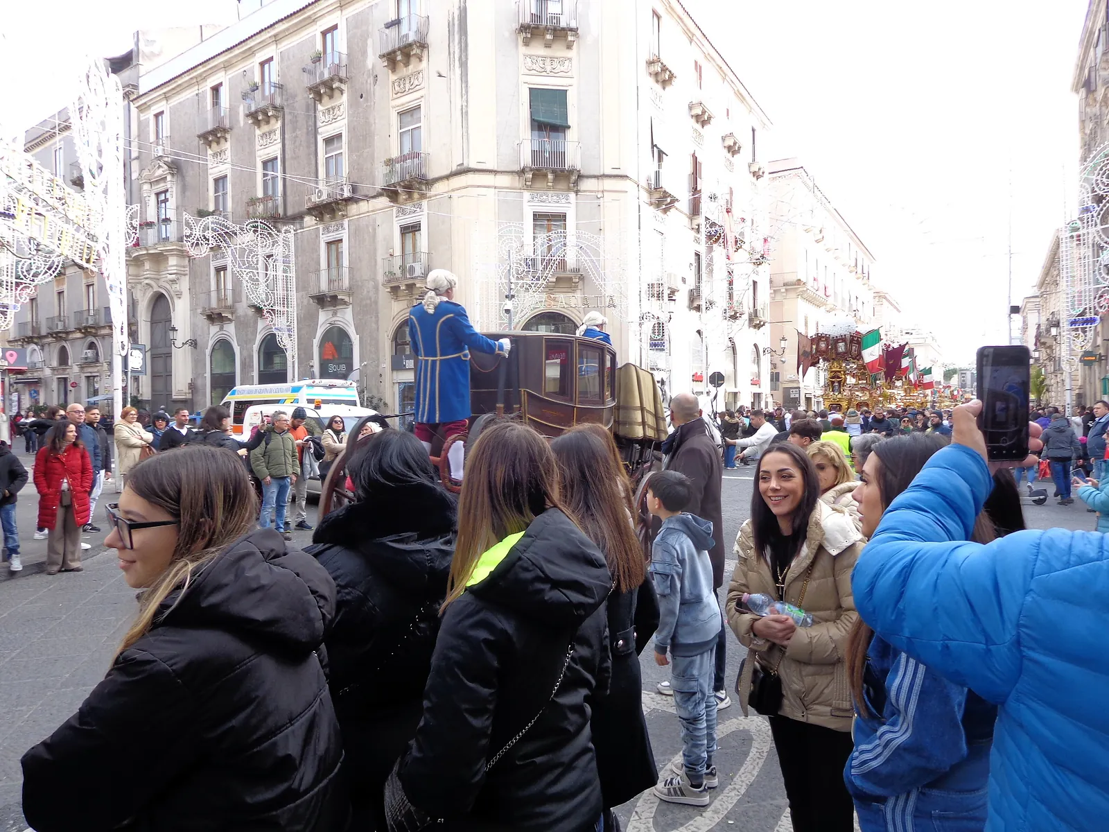 A dark antique carriage with large spoked wheels standing in a wide street, a coachman in a blue and gold livery coat and white periwig on the box. Strings of white filigree light frames run overhead between the buildings, a crowd in winter coats watches from the pavement, and further down the street a tall gilded tower stands among small Italian flags.