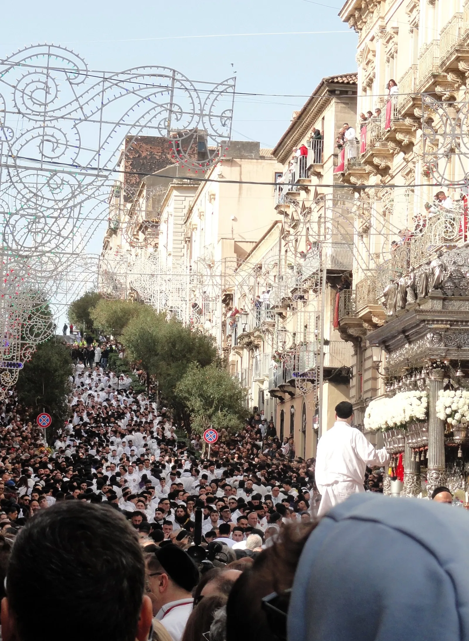 Looking up a street so full of people that no road surface is visible: hundreds in white smocks and black caps among others in dark coats, with a line of them continuing up a stepped stretch beyond. Arches of white filigree lights hang above, spectators fill every balcony, and at the right edge the silver shrine stands level with the upper floors, banked with white flowers.