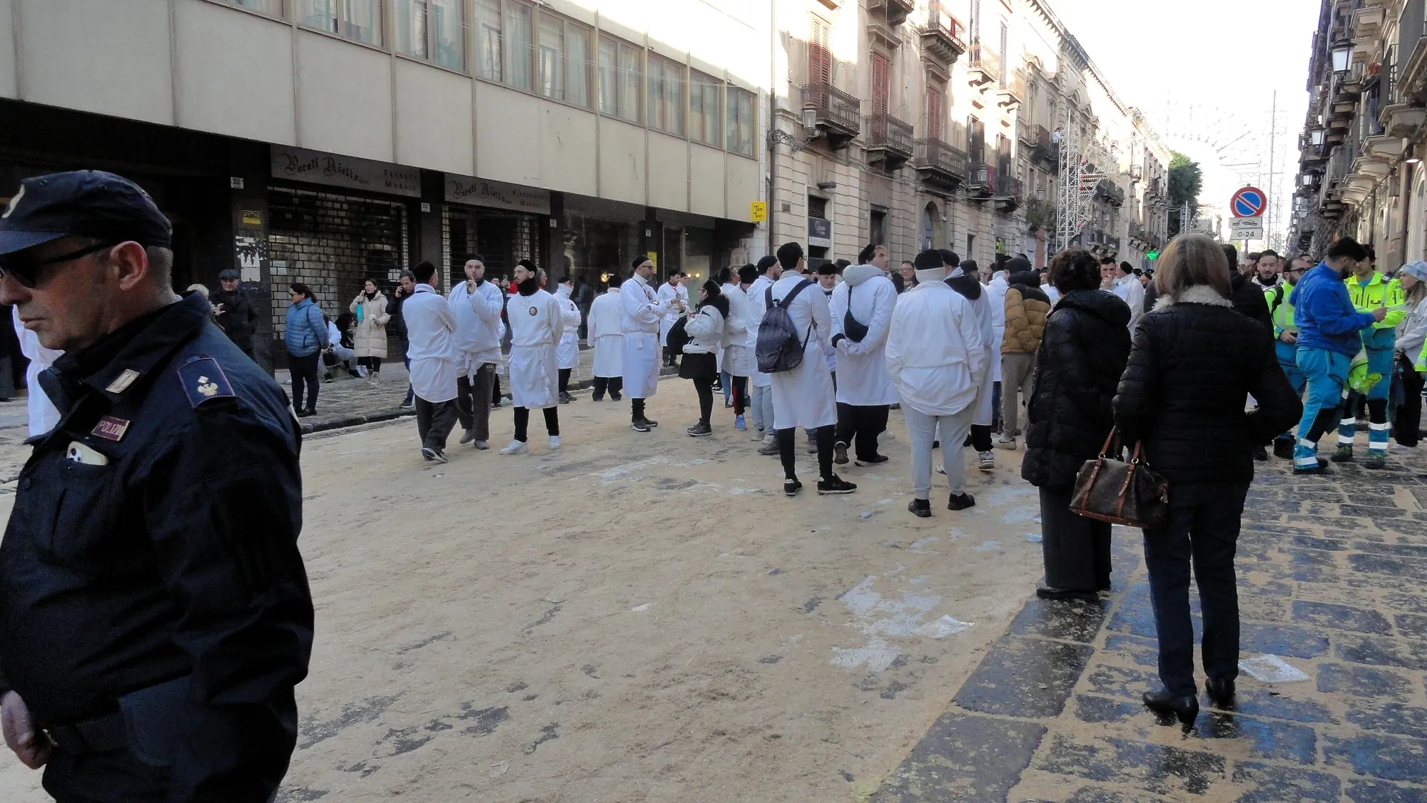 A wide city street with men in white cotton smocks standing in loose groups down the middle of it, some with a small emblem on the back. The roadway is covered in an even pale layer underfoot. A policeman stands in the left foreground, ambulance crew in high-visibility jackets wait at the right, and an arch of unlit light frames spans the street in the distance.