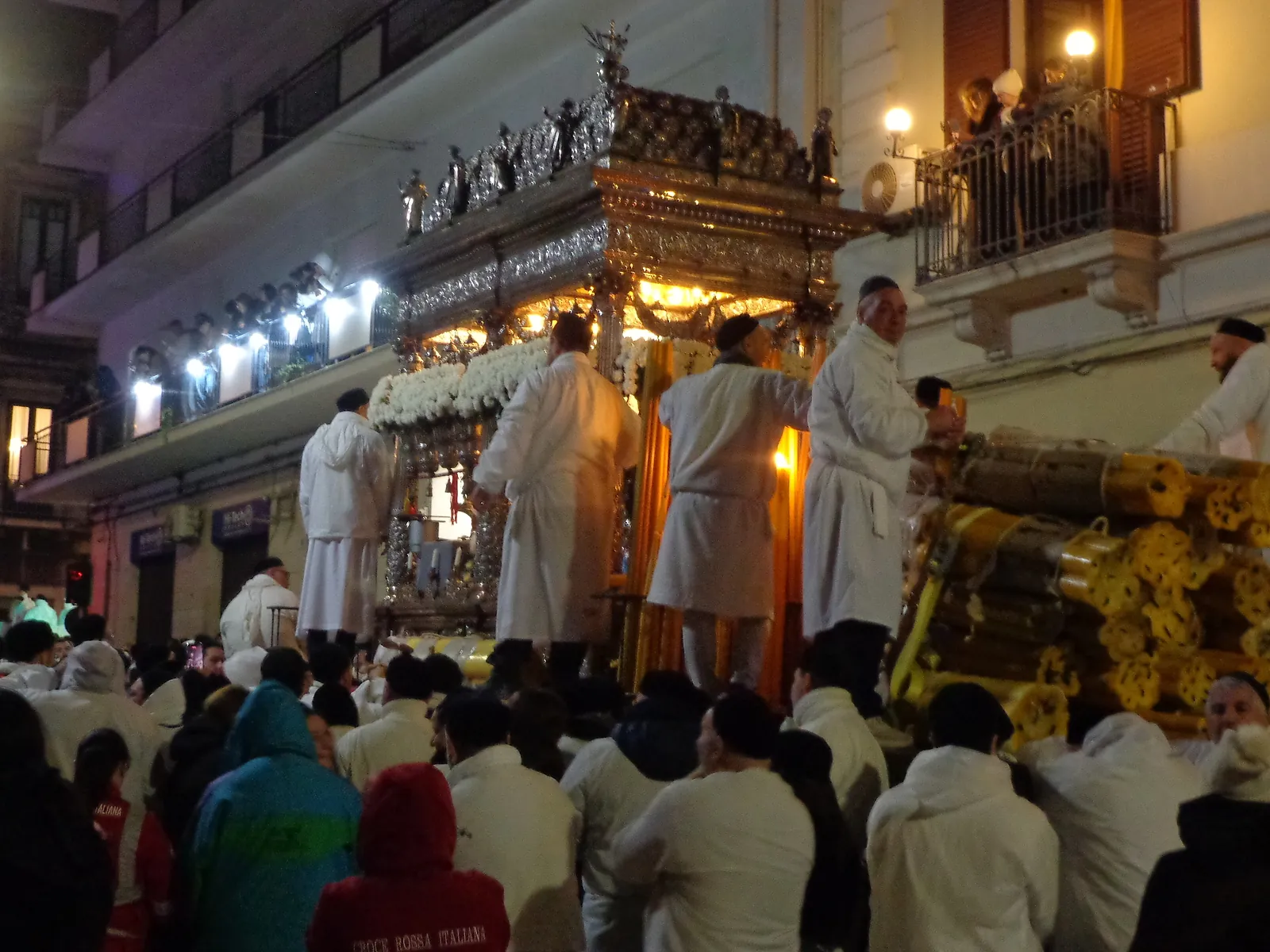 A silver and gilt shrine at night, its canopy roof crowded with small standing figures and its columns wound with white carnations, lit from within. Men in white cotton smocks and black skullcaps stand around it, one reaching up to the platform. At the right a bundle of long gilded poles with ornamented ends leans against the crowd, and people watch from a balcony above.