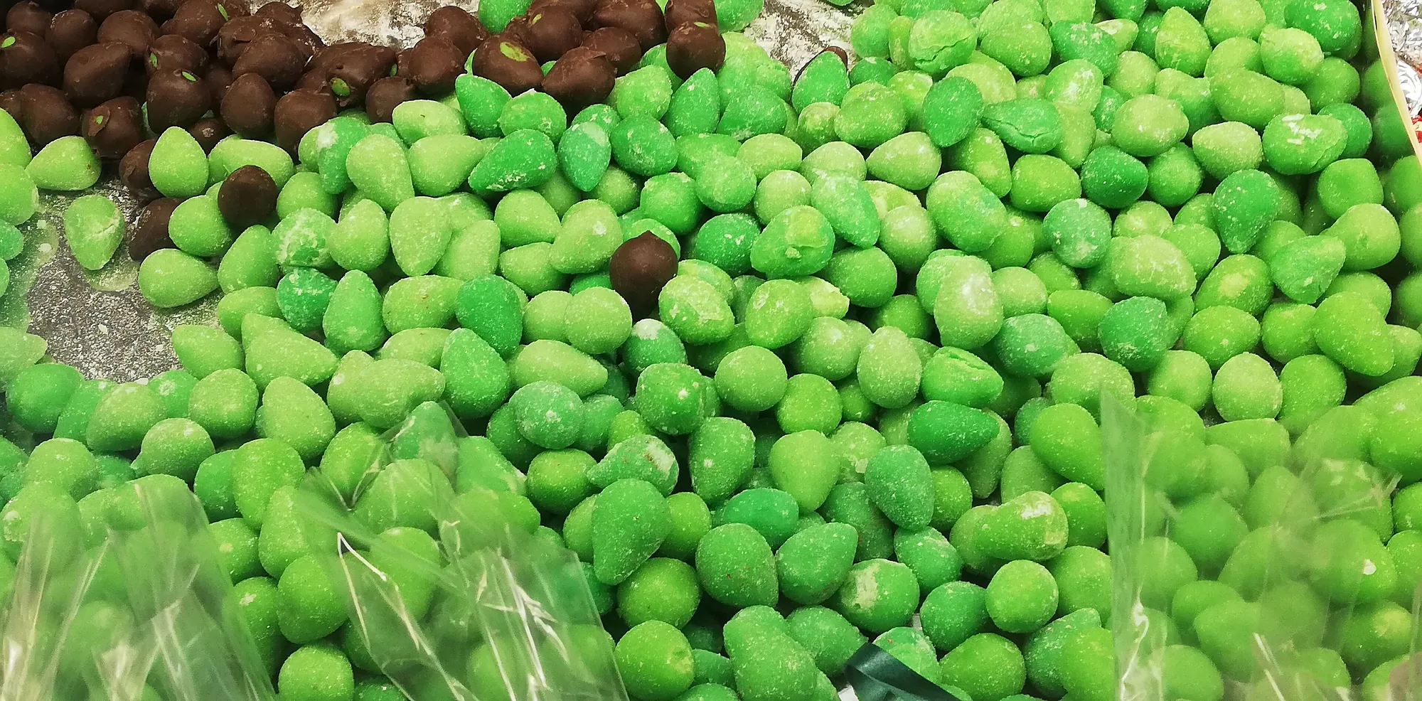 A shop display heaped with small bright green marzipan olives, dusted and slightly irregular, with a smaller heap of chocolate-coated ones behind them and clear cellophane bags at the front edge.
