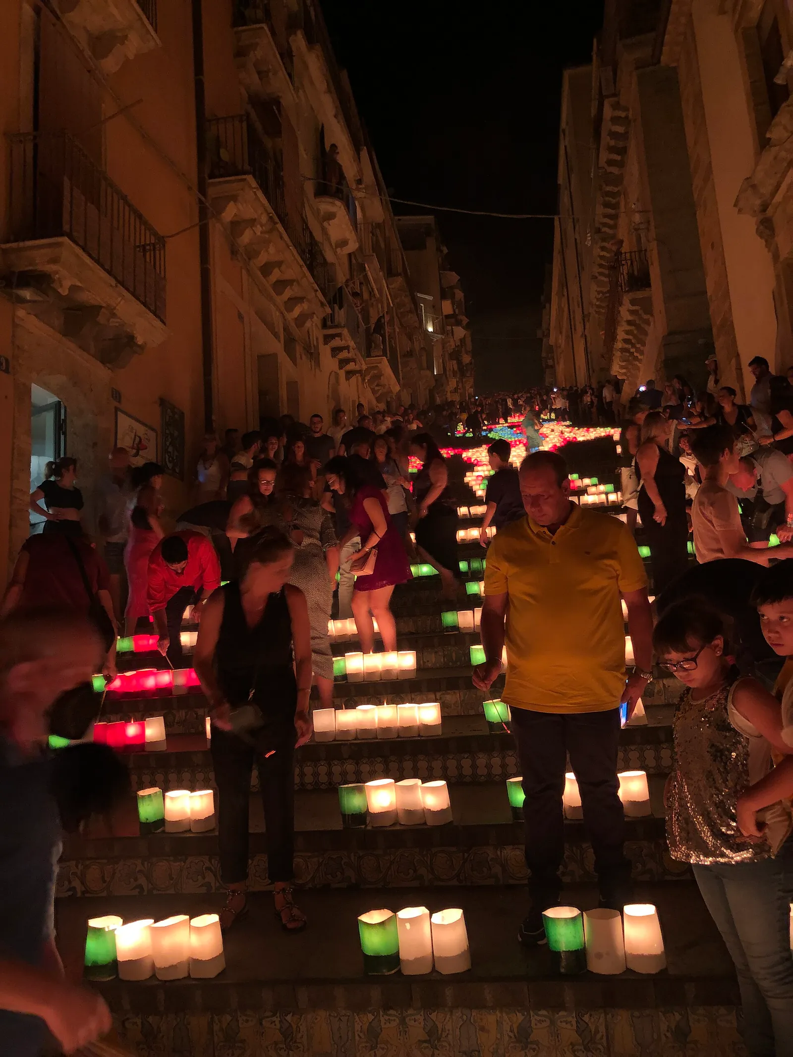 A long flight of steps at night, seen from partway up. On every tread stand rows of small glowing lamps — white, green, pink and red — set out in a pattern that runs away up the staircase into the dark. People stand and walk among them on the steps; the patterned maiolica facing of the risers shows in the lamplight, and balconied houses rise steeply on both sides.