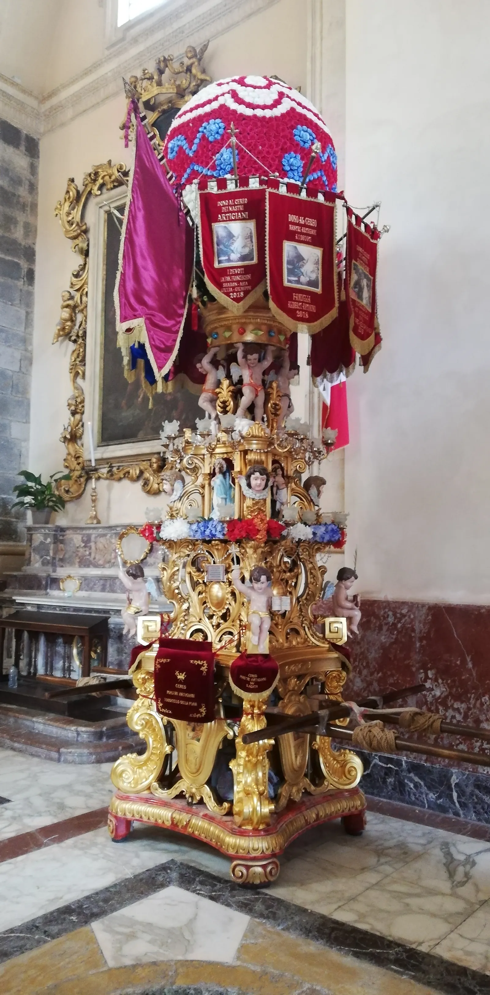 A carved and gilded wooden tower about three men high, standing on the marble floor of a cathedral in front of a side altar. It is built in tiers of scrollwork with painted putti and angels sitting on the cornices and a small glazed niche of figures at mid-height; above it hangs a crowned canopy with dark red embroidered banners, and on top sits a domed cushion of red, white and blue flowers. Two long carrying beams are slotted through the red-and-gold base, one with a coil of rope on it.