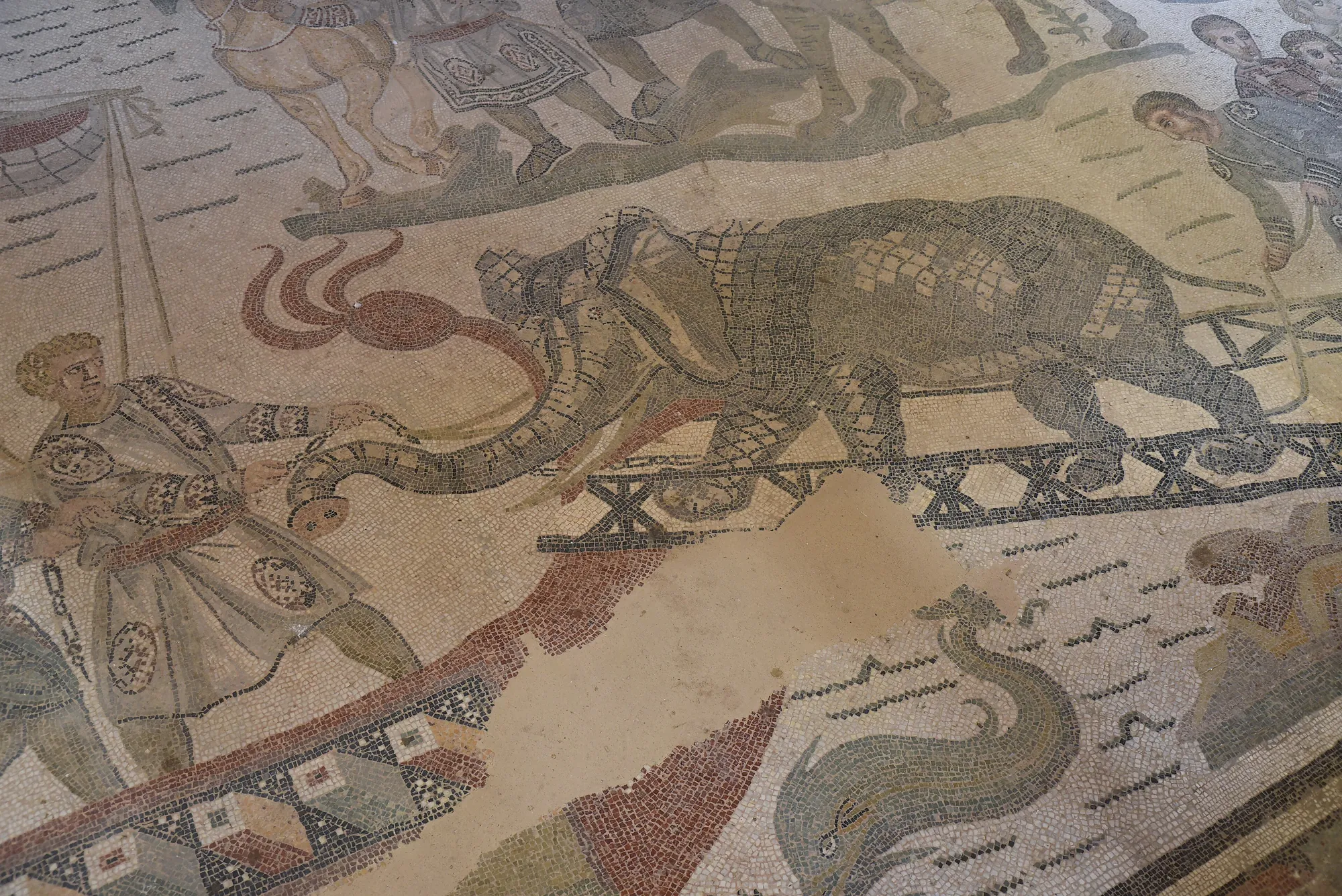 A fourth-century floor mosaic: an elephant in a body harness walking up a plank gangway towards a ship, a handler in a short tunic at its head holding the rope, more figures and animals above, and a red-and-cream boat hull below, all in grey, ochre and brick-red tesserae on a cream ground with bare patches of restored plaster.