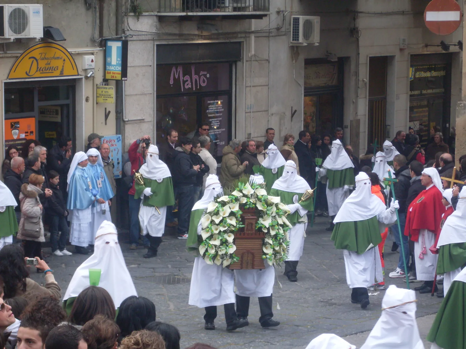 A town street with men in long white robes and pointed white hoods with eye slits, wearing green shoulder capes, carrying a bier heaped with white arum lilies around a small carved model of a building. Others among them wear pale blue capes and deep red capes, and onlookers stand along the shopfronts behind.