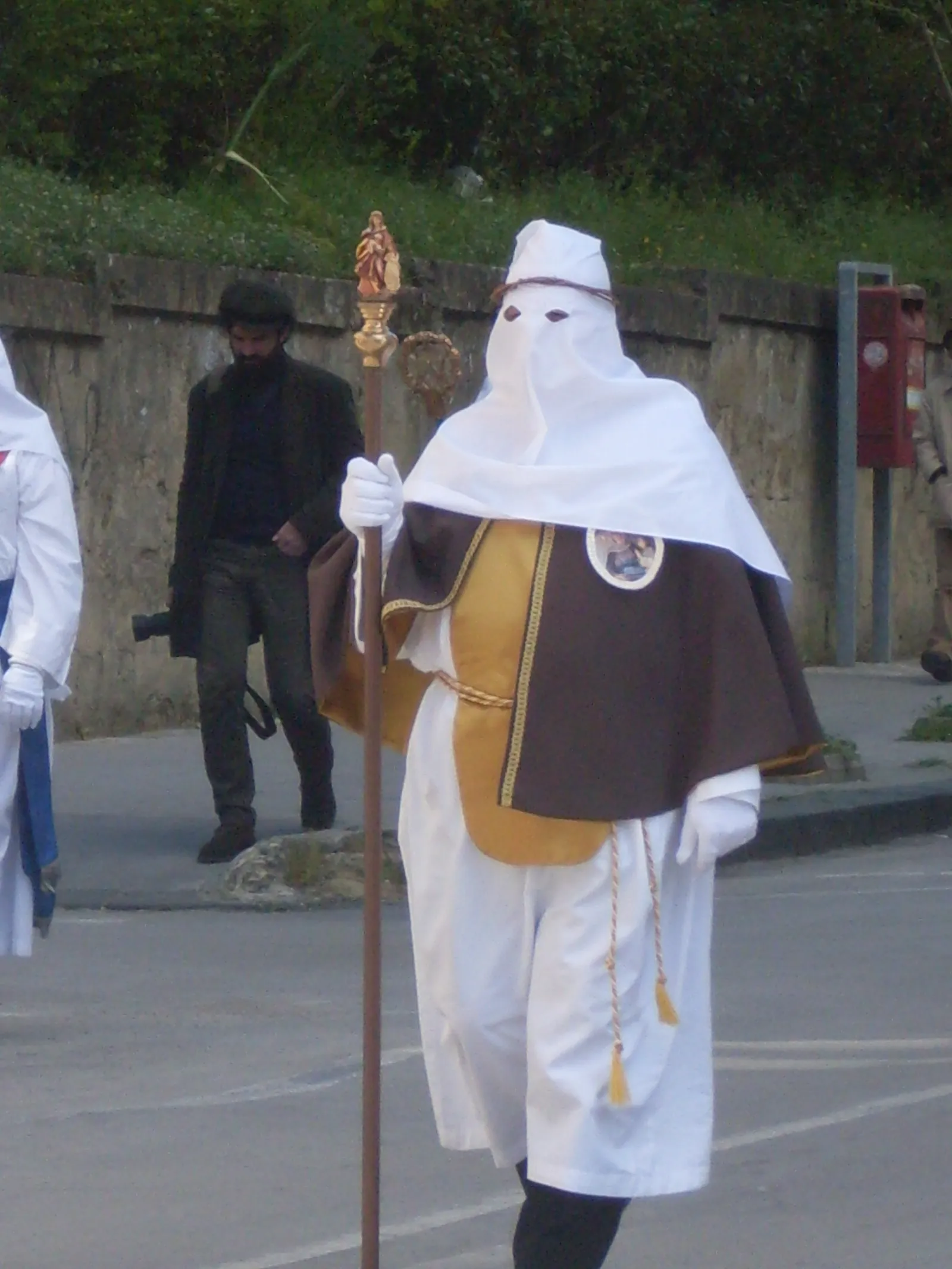 One hooded man walking alone on a road: a tall pointed white hood with narrow eye slits and a twisted band of withy around it, a white robe, and over it a brown and gold shoulder cape bearing an oval painted medallion. He wears white gloves and carries a tall staff topped with a small gilded figure. A second hooded figure is partly visible at the left edge.