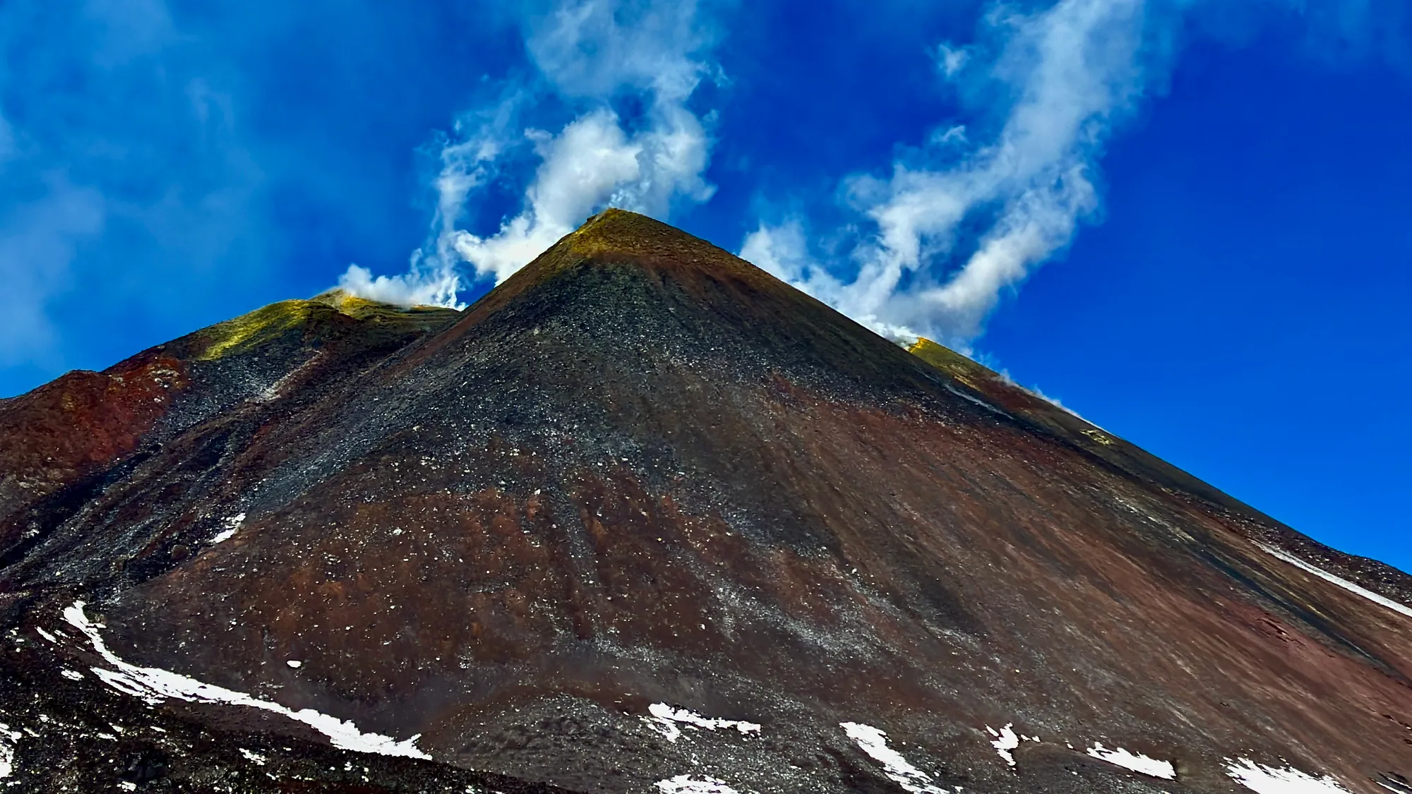 A steep, near-triangular volcanic cone seen from below against a deep blue sky. Its slopes are dark brown, black and rust-red loose cinder, gullied by erosion. Bright yellow-green sulphur staining marks the crest and both upper shoulders, a white plume streams off the summit, and patches of snow lie at the foot of the slope.