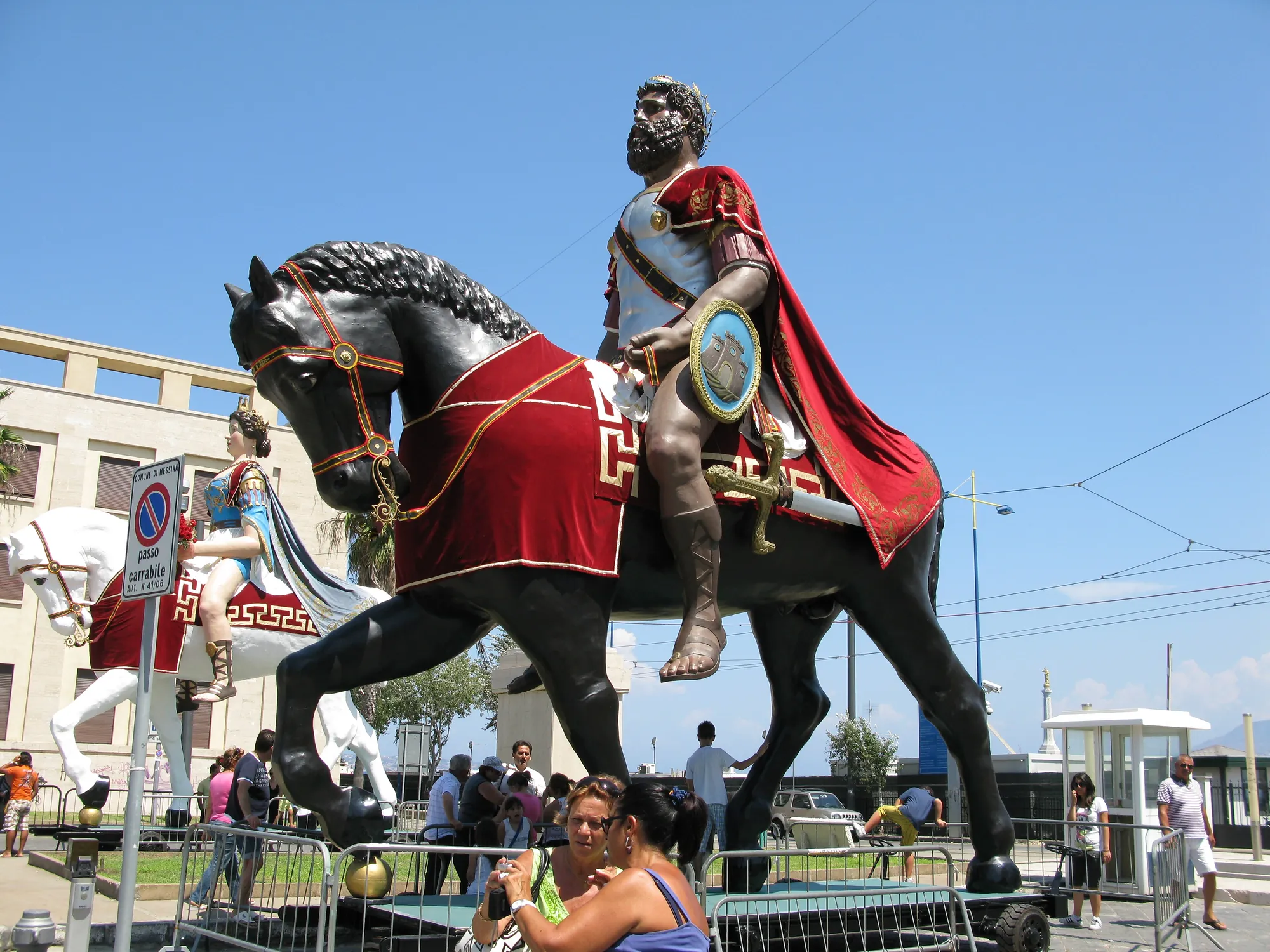 Two large painted statues on low wheeled platforms in a sunlit street: a dark-skinned bearded warrior in Roman armour and a long red cloak astride a black horse, and behind him a crowned pale-skinned woman in blue and white on a white horse. Metal crowd barriers stand around them and onlookers sit in the shade.