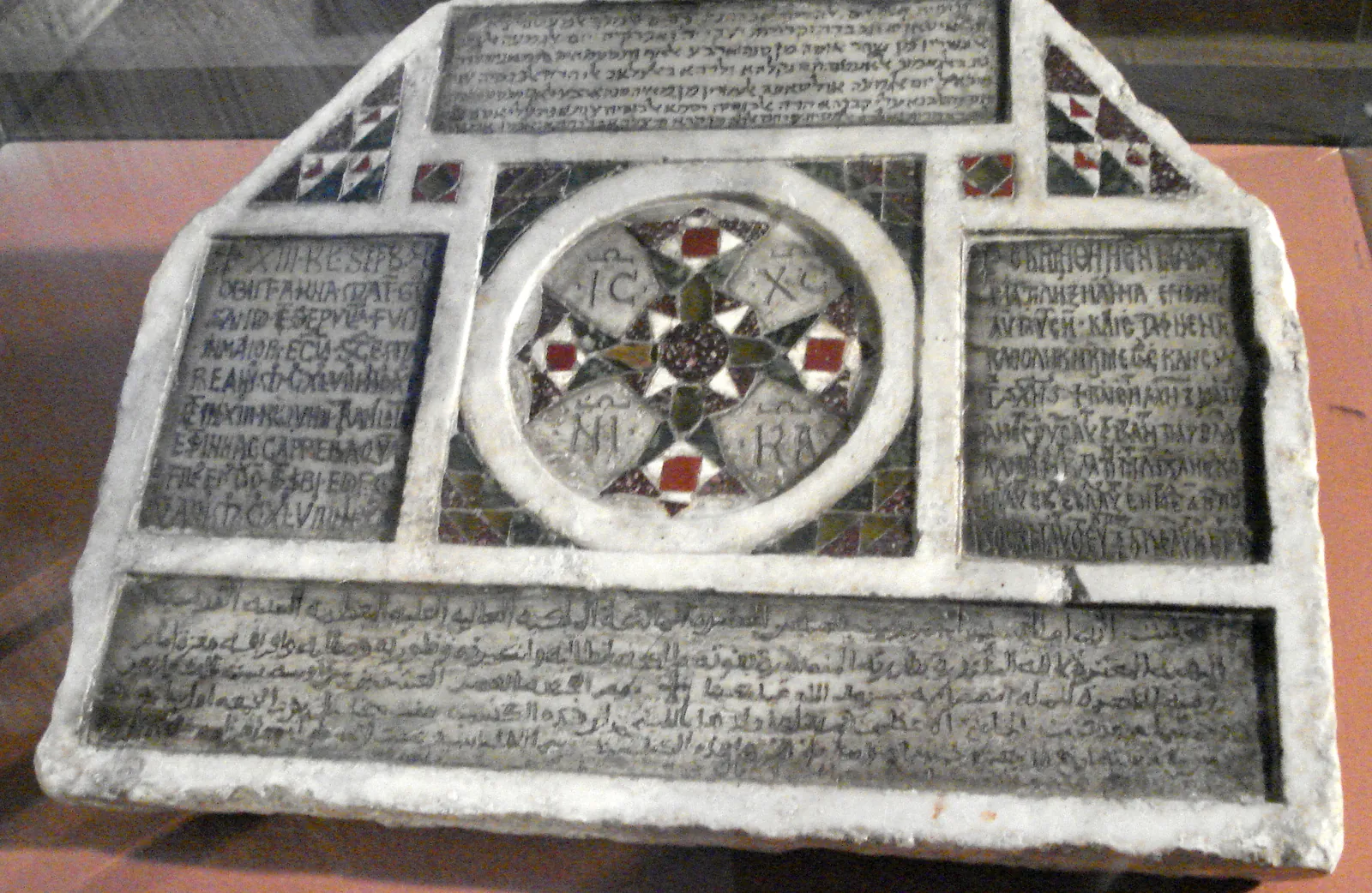 A trapezoidal white marble slab standing in a museum case. Four sunken inscribed panels surround a central roundel: Hebrew letters in a band across the top, Latin at upper left, Greek at upper right, and Arabic running the full width of the lower band. The roundel holds an eight-pointed cross inlaid in red, green and white stone, lettered IC XC NI KA in its four quarters.