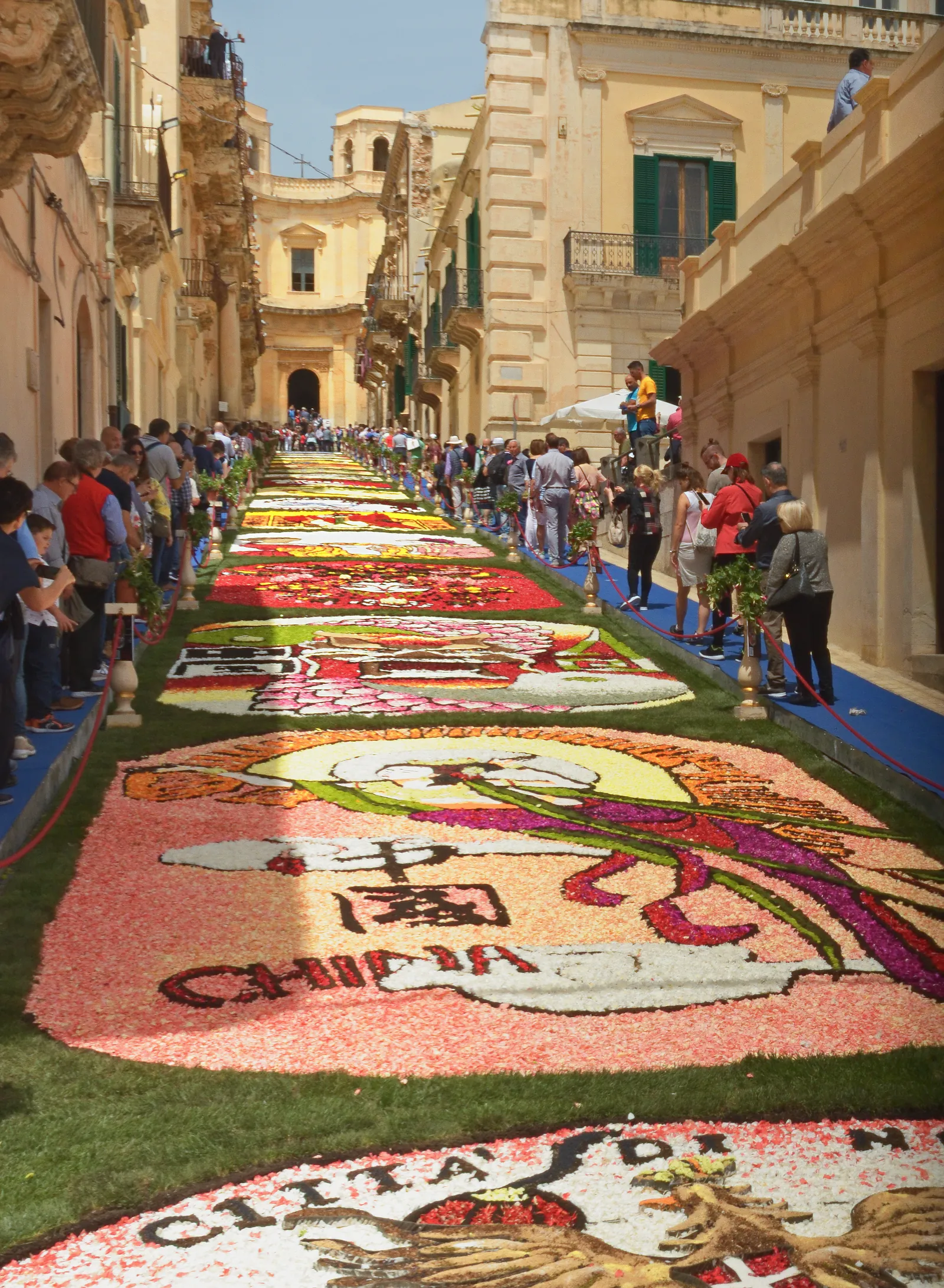 A sloping street seen from its lower end, filled from wall to wall with panels of coloured flower petals laid on the paving — pinks, reds, greens and cream, framed in strips of green. Spectators line both sides behind red rope barriers; carved stone balconies overhang from the left and a domed church closes the view at the top of the slope.