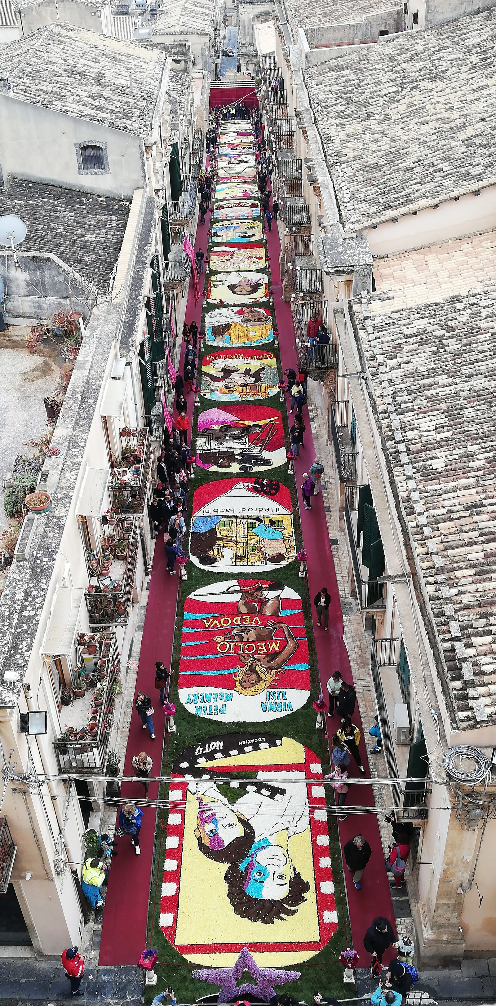 Looking straight down onto a narrow street from a bell tower: a long strip of bright flower-petal panels running between two rows of tiled rooftops and terraces, edged with a deep red border, with small figures walking the margins and washing lines and plant pots on the balconies to either side.