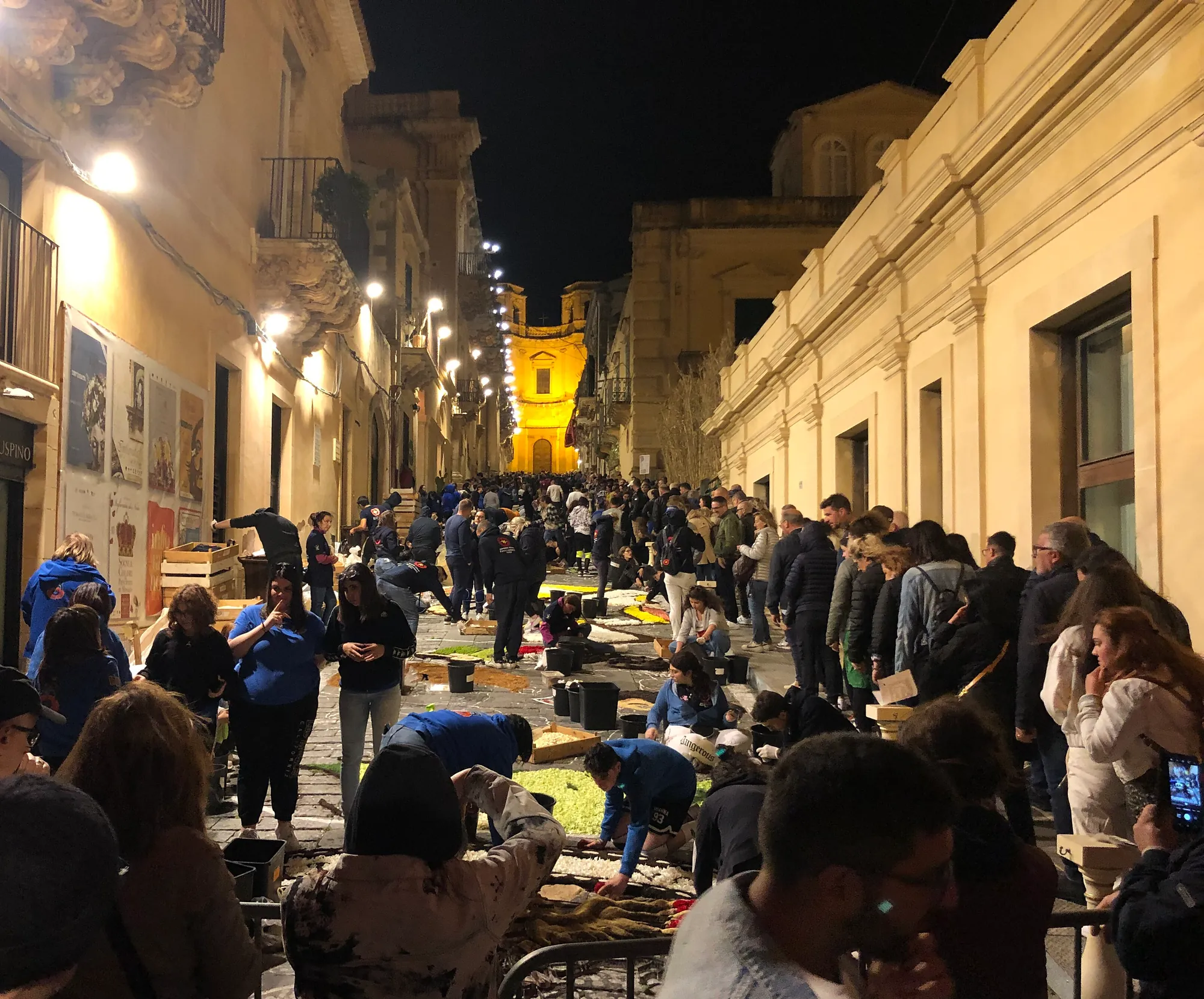 The same street at night, lit by street lamps, with a church glowing gold at the top of the slope. Dozens of people in blue team shirts kneel on the paving laying handfuls of petals, with wooden crates, trays and black buckets around them. Onlookers stand in rows along both walls behind barriers.