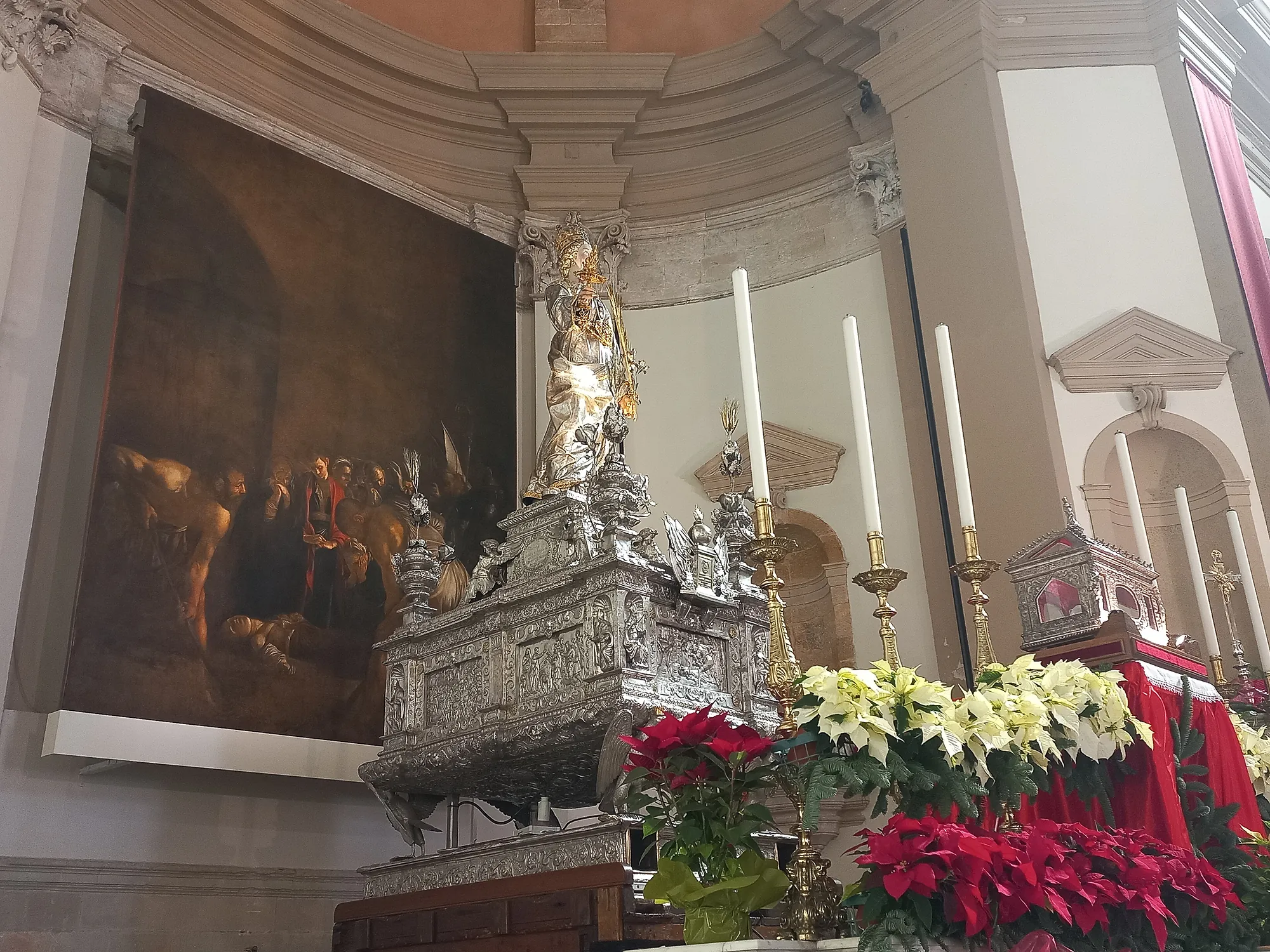 A church interior seen from below. On the left hangs a very large dark canvas of a burial scene, two muscular gravediggers in the foreground and a crowd of mourners behind. To the right of it a silver figure of a woman stands on top of an ornate silver chest worked in relief, with tall white altar candles behind and banks of red and white poinsettias below.
