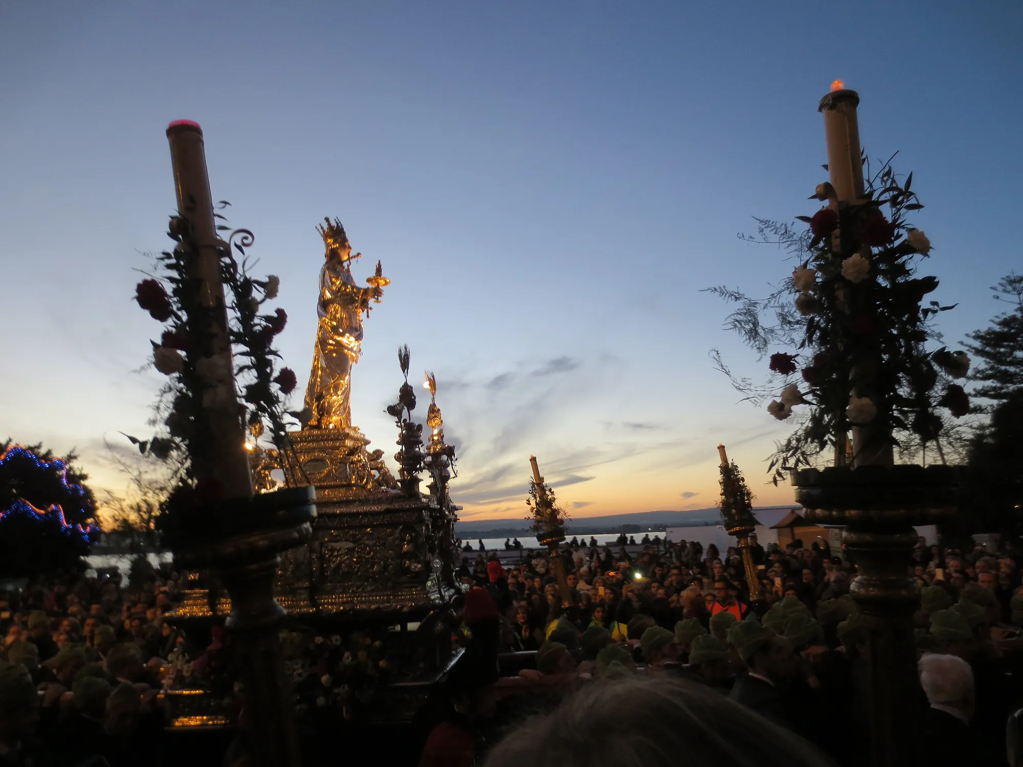 Dusk over water and a low coastline. A gilded and silver figure of a crowned woman stands on a heavily worked silver bier, flanked by tall thick candles wound with dark red and white flowers and sprays of foliage. Below and around, a dense crowd of people in dark coats, most of them wearing soft green caps.