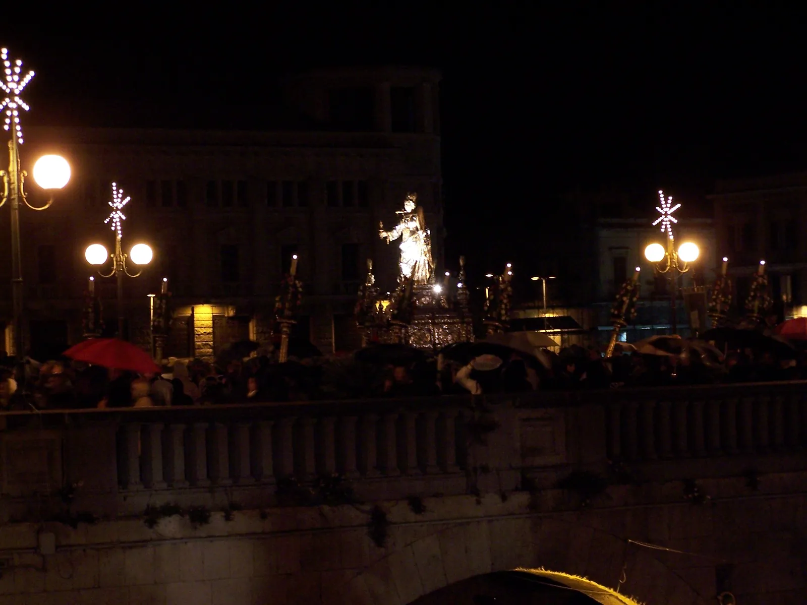 Night, and raining. The lit silver figure is carried along a stone balustraded bridge among a crowd holding black umbrellas, with tall flower-wound candle-poles around her and star-shaped decorative lamps on the street lights. A pale building front stands behind and the arch of the bridge is visible below.