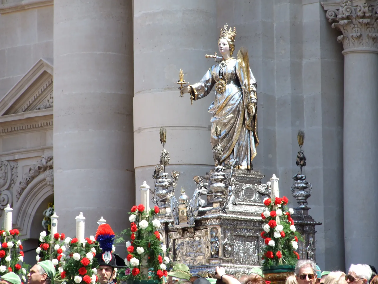 Daylight. The silver figure, crowned and holding a lamp and a palm, stands on an elaborately worked silver bier banked with red and white carnations, in front of enormous plain fluted stone columns and a carved baroque wall. Bearers in green caps and a carabiniere in a plumed bicorn hat stand below.