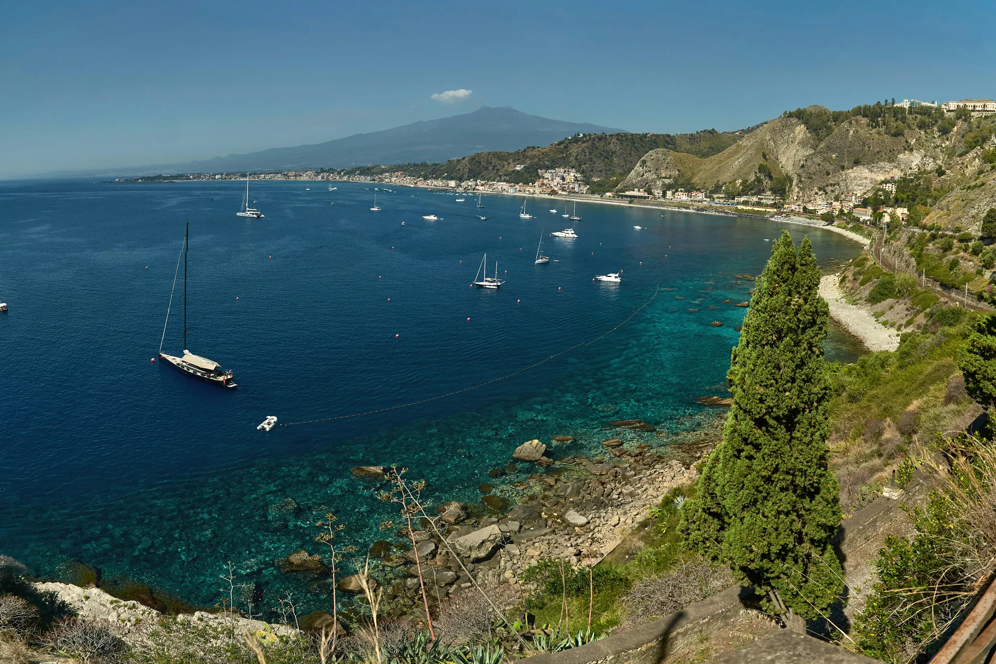 Looking down from a cliff path, past agaves and a tall cypress, onto a wide blue bay. Turquoise shallows and dark reef fringe the near shore, a dark-hulled yacht and half a dozen white ones lie at anchor, a long beach across the water is backed by a strip of low town buildings, and behind them a broad volcano rises to a cloud-wrapped summit.