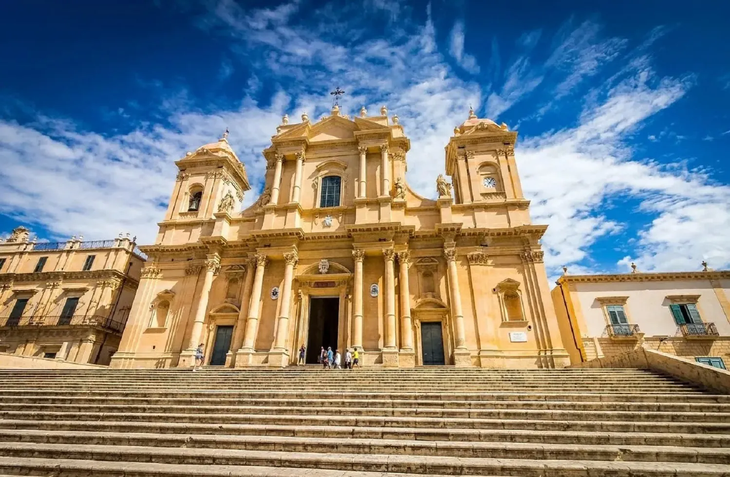 A honey-coloured Baroque church front seen straight on under a blue sky. Three arched doorways at ground level are framed by pairs of tall Corinthian columns; a square bell tower capped with a small dome stands at each end, the right one carrying a clock face. Statues stand in niches and along the parapets, and a broad flight of steps runs up to the doors.