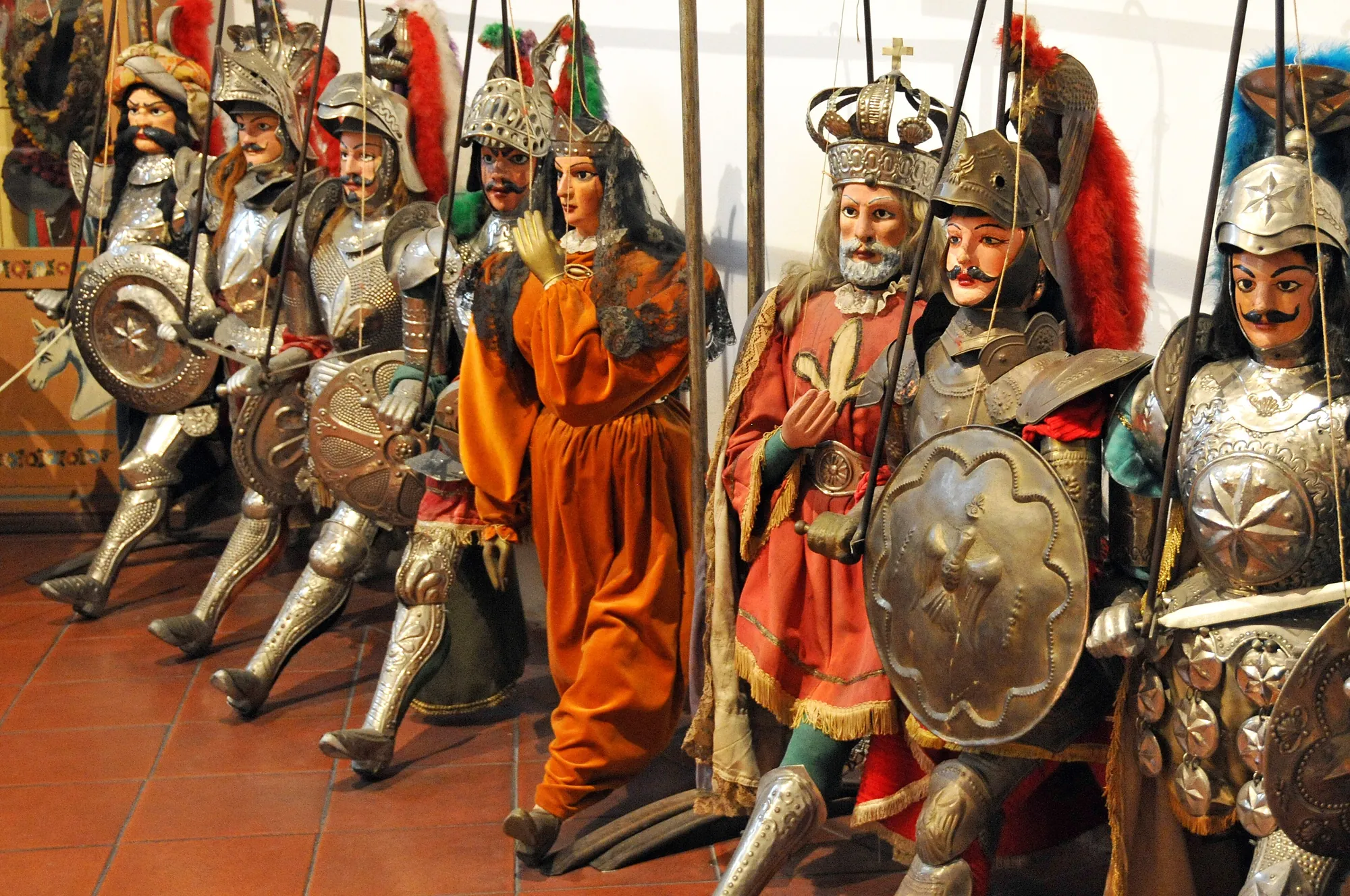 Six or seven marionettes standing in a row, seen from just above knee height. Knights in embossed silver and brass plate armour with hinged leg and arm pieces, visored helmets with red, green and blue feather plumes, and large engraved round and kite shields held forward; among them a female puppet in an orange gown with a black lace shawl, and on the right a crowned, white-bearded king in a red and gold robe. Control rods and strings run up out of the top of the frame.