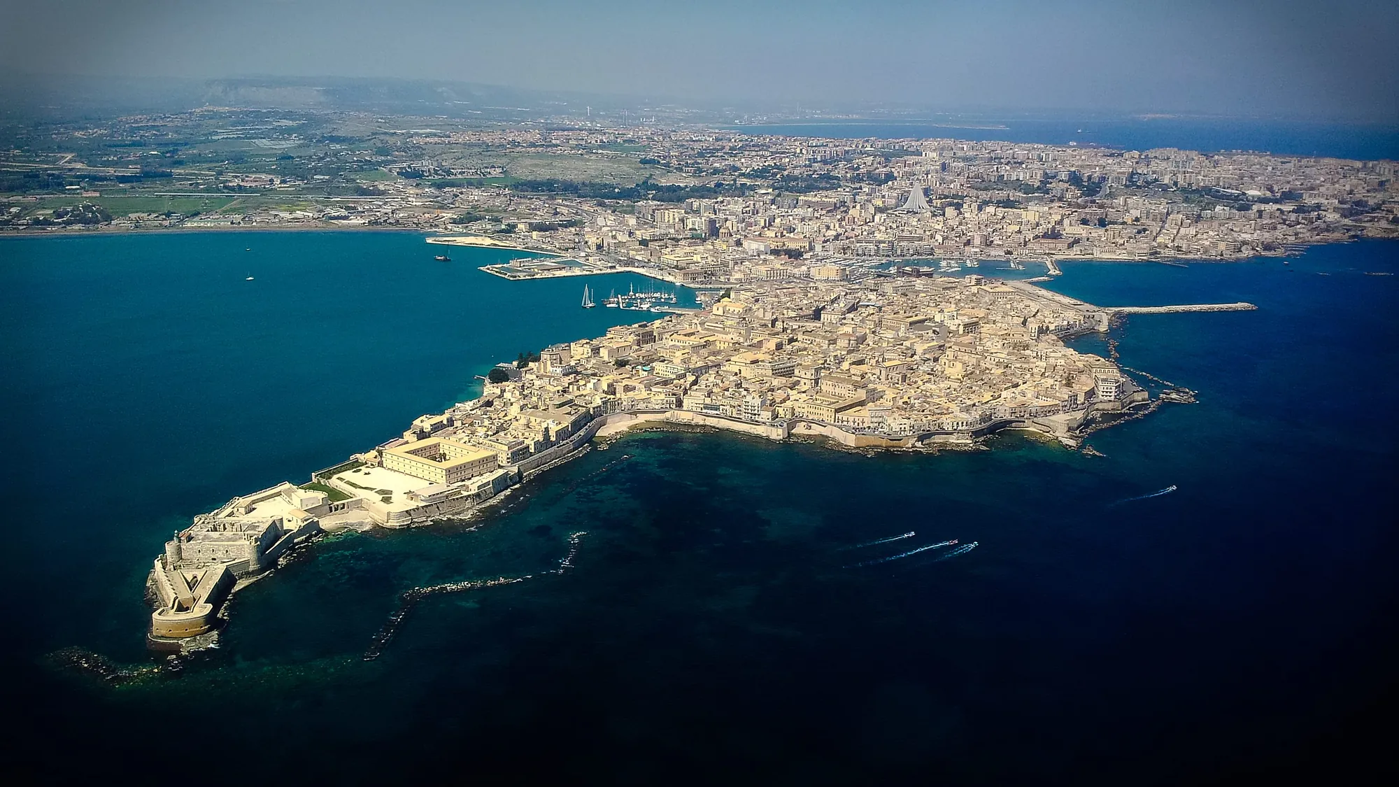 A high aerial view of a long narrow island covered edge to edge with pale flat-roofed buildings, joined at one end to a mainland city that stretches to the horizon. A square fortress with a round tower stands on the island's far point. Turquoise water and a marina full of sailing boats lie on one side, darker open sea on the other.