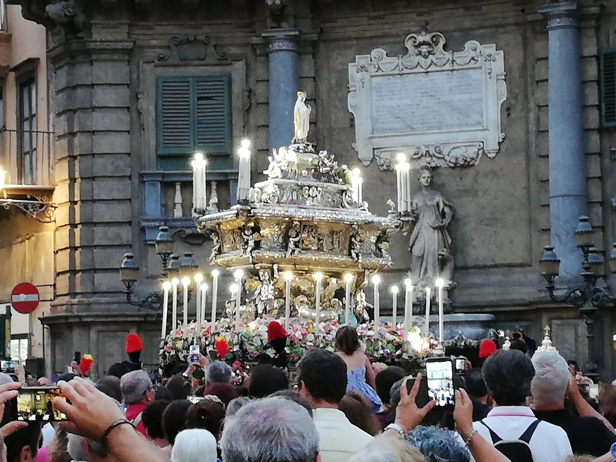 Dusk in a city street. A tiered silver and gilt reliquary ark stands raised above a dense crowd, ringed by tall lit candles and banked with flowers, a small figure on its summit. Behind it rises the grey stone of a baroque corner building carrying a carved marble inscription tablet and a statue in a niche. Many in the crowd are holding up phones.