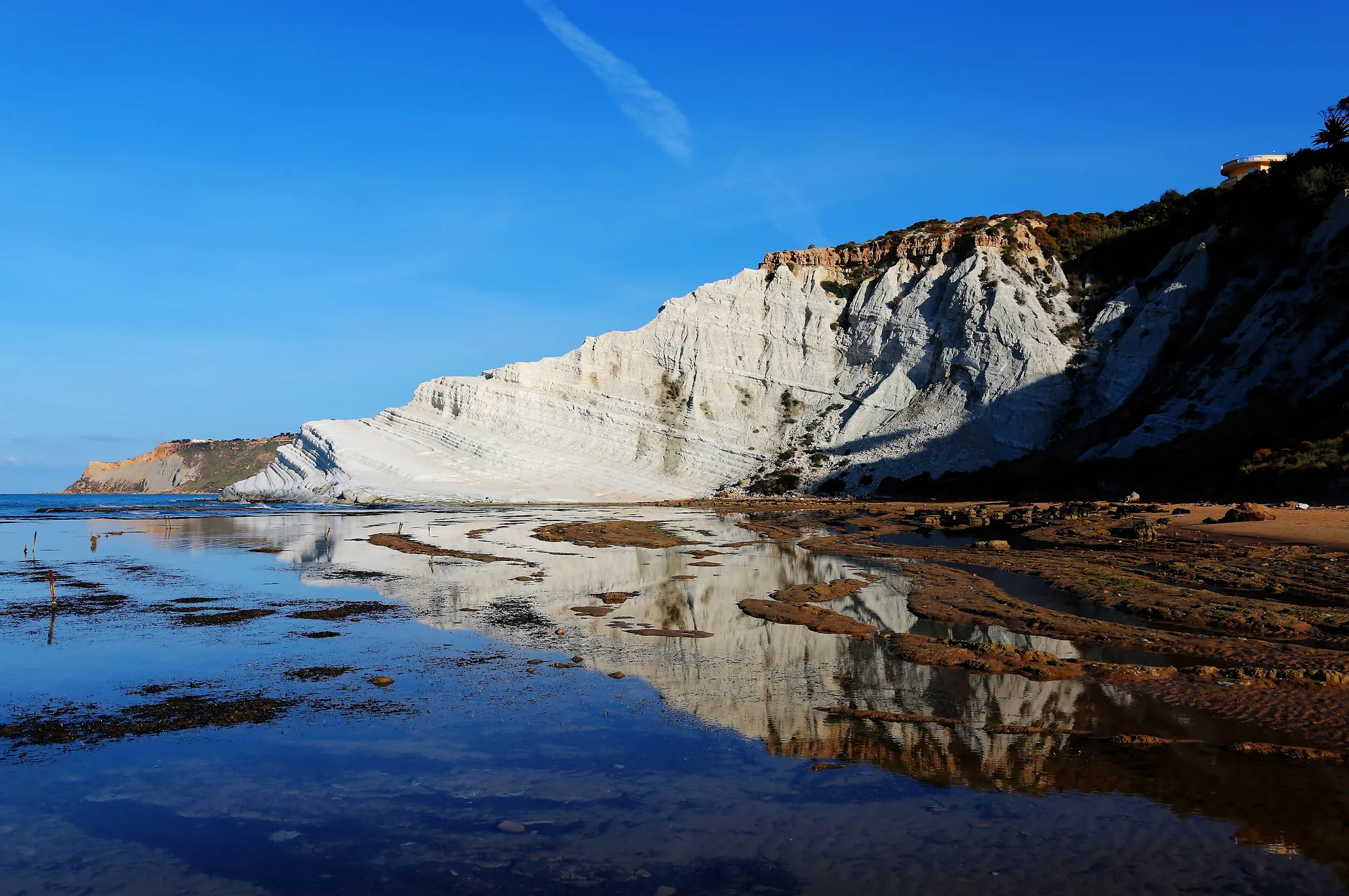 A white marl headland whose horizontal beds step down like a staircase into the sea, seen from the beach at its foot in low warm sun, with dark scrub and a band of rust-coloured rock along its top edge and the whole cliff mirrored in a thin sheet of water over the tidal flat in the foreground.