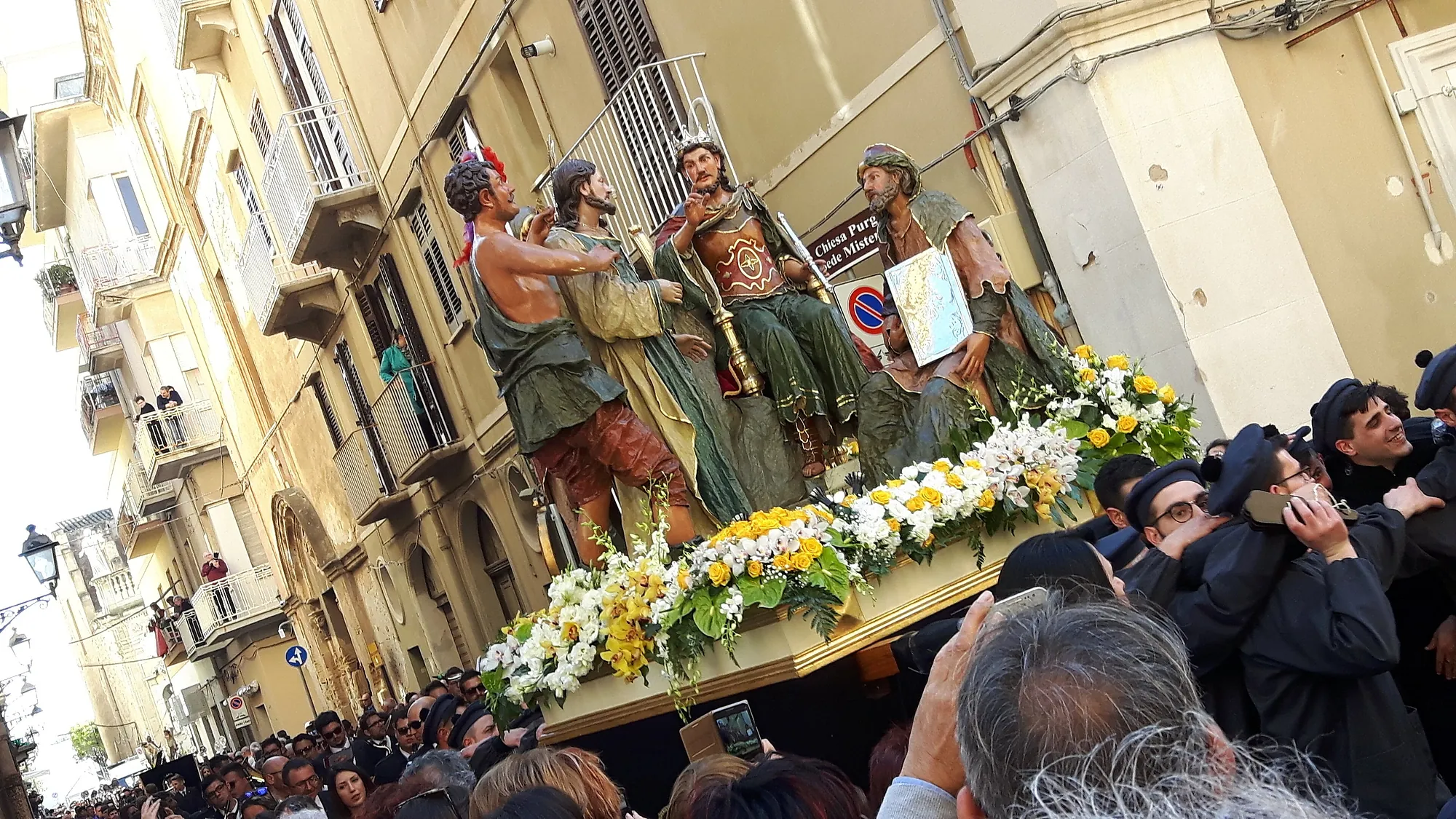 A carved and painted group of four life-size figures on a float carried at shoulder height down a narrow street: a soldier with a feathered cap, Christ bound with a rope, a seated man in a green robe holding a staff, and a robed figure holding a written scroll. The float's edge is banked with yellow and white flowers. Men in dark suits are pressed beneath it, and spectators fill the balconies above.