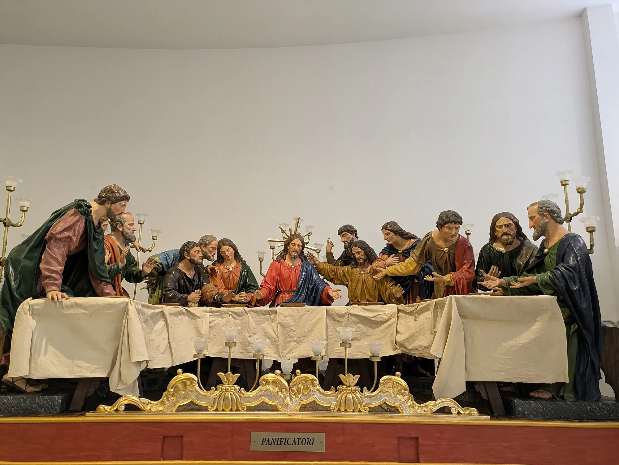 A life-size polychrome sculpture group of the Last Supper standing in a white-walled room: thirteen figures in coloured robes behind a long table draped in cream cloth, Christ at the centre with arms open, candelabra at either end. A gilded scrollwork crest is fixed to the front of the platform, and a plaque below it reads PANIFICATORI.